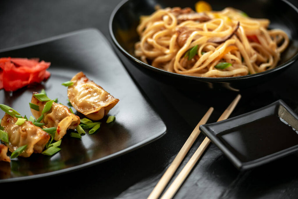 Asiatisches Essen auf dunklem Hintergrund auf schwarzem Teller. Japanische gebratene Gyozo-Knödel und Udonnudeln. - Foto, Bild