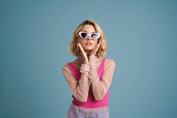 Young white woman in sunglasses gesturing and looking at camera isolated over blue wall - Foto, afbeelding