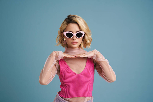 Young white woman in sunglasses gesturing and looking at camera isolated over blue wall - Фото, зображення