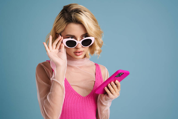 Young white woman in sunglasses using mobile phone isolated over blue wall - 写真・画像