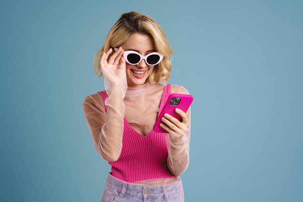 Young white woman in sunglasses smiling while using mobile phone isolated over blue wall - Foto, afbeelding