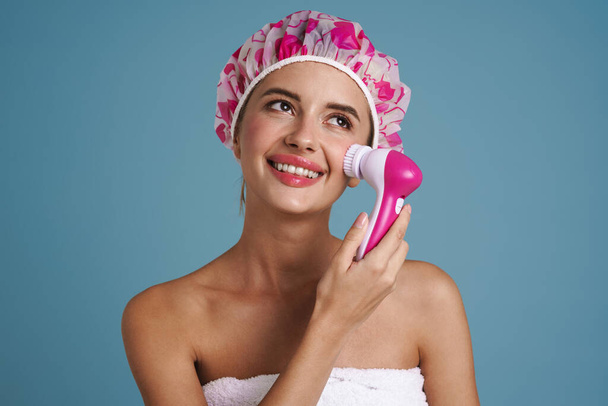 Young woman wearing shower cap smiling while using facial massager isolated over blue wall - Фото, изображение