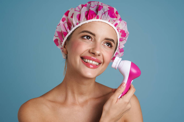 Hemdlose Frau mit Duschkappe lächelt, während sie Gesichtsmassagegerät über blauer Wand isoliert benutzt - Foto, Bild