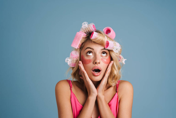 Young woman with curlers and eye patches expressing surprise isolated over blue wall - Photo, image