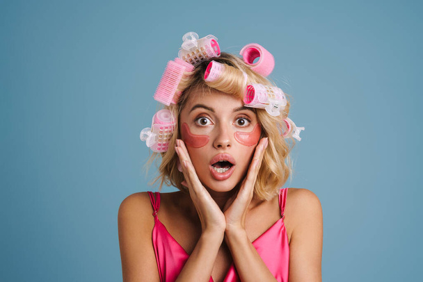 Young woman with curlers and eye patches expressing surprise isolated over blue wall - Foto, Imagem