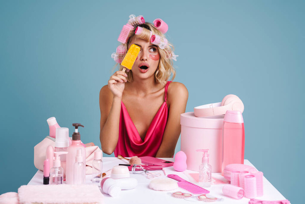 Surprised woman holding ice cream while sitting at table with beauty products isolated over blue wall - Φωτογραφία, εικόνα