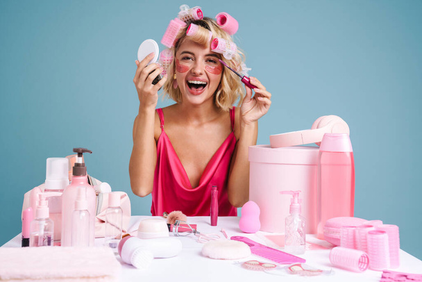 Young woman with curlers doing makeup while sitting at table isolated over blue wall - Foto, imagen