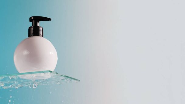Mockup of a white dispenser with a cosmetic product on glass shelf and falling down drops on blue background closeup. Cosmetic product for body washing - 写真・画像