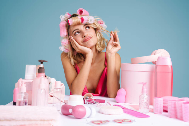 Young woman smiling while sitting at table with beauty products isolated over blue wall - 写真・画像
