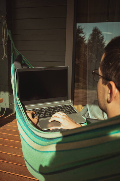 Een comfortabele plek om thuis te werken. Een man zit en werkt in een hangmat bij een landhuis op een zonnige dag. - Foto, afbeelding