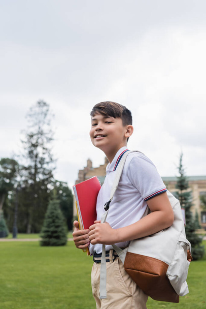 Smiling asian schoolboy holding notebook and backpack outdoors  - Фото, изображение