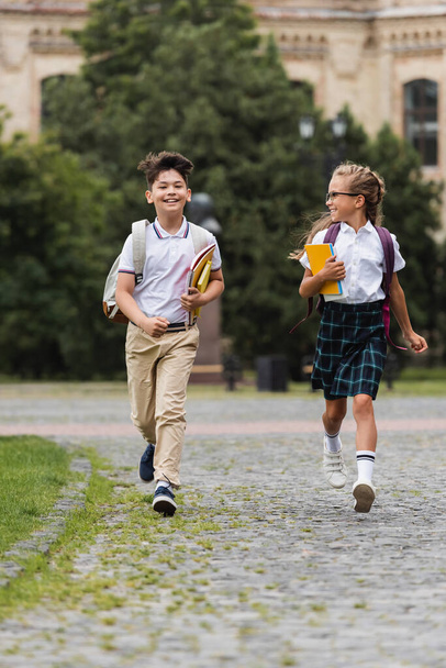 Positive multiethnic pupils with backpacks running on walkway outdoors  - Photo, Image