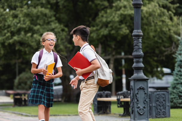 Asian schoolboy with backpack talking to cheerful friend in eyeglasses in park  - Fotó, kép