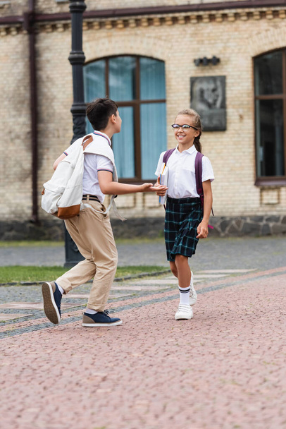 Asian schoolboy with backpack talking to cheerful friend with notebooks outdoors  - Foto, Imagen