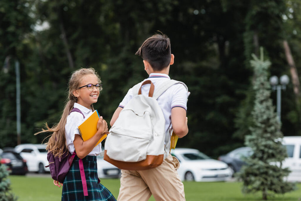 Positive schoolgirl in eyeglasses holding notebooks near classmate outdoors  - Photo, Image