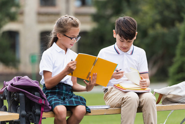 Preteen multiethnic schoolkids holding notebooks on bench outdoors  - Zdjęcie, obraz