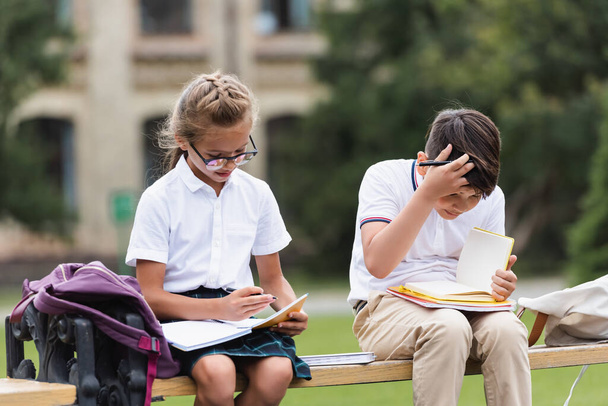 Interraciale schoolkinderen kijken naar notitieboekjes op bank buiten  - Foto, afbeelding