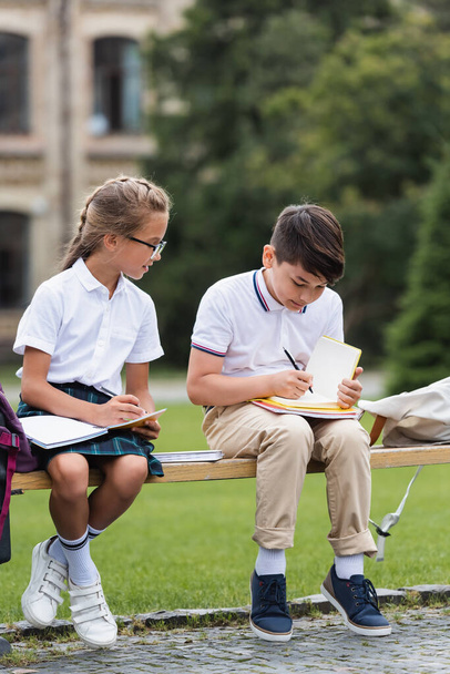 Asian schoolboy writing on notebook near friend on bench outdoors  - Фото, изображение