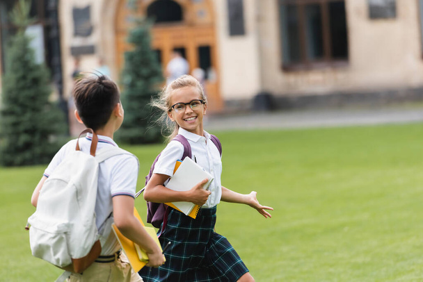 Mosolygó iskolás lány jegyzetfüzetekkel sétál az osztálytársa mellett a gyepen a parkban  - Fotó, kép
