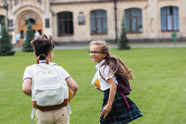 Positive schoolkid in eyeglasses running near asian friend with backpack on lawn  - Φωτογραφία, εικόνα