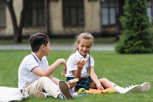 Positive schoolgirl holding sandwich near asian classmate on lawn in park  - Photo, Image