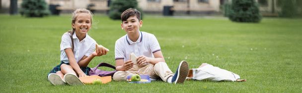 Pozytywne międzyrasowe dzieci trzymające kanapki w pobliżu plecaków na trawie w parku, transparent  - Zdjęcie, obraz