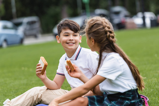Smiling asian schoolboy holding sandwich near blurred friend on lawn in park  - Photo, Image