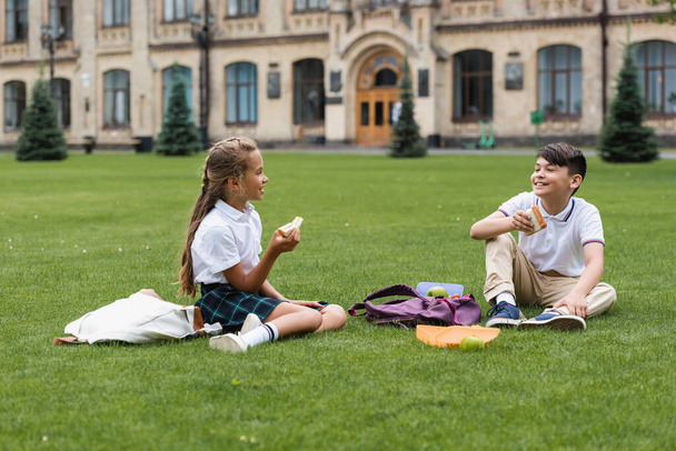 Multiethnic schoolkids holding sandwiches and talking on grass in park  - Фото, зображення