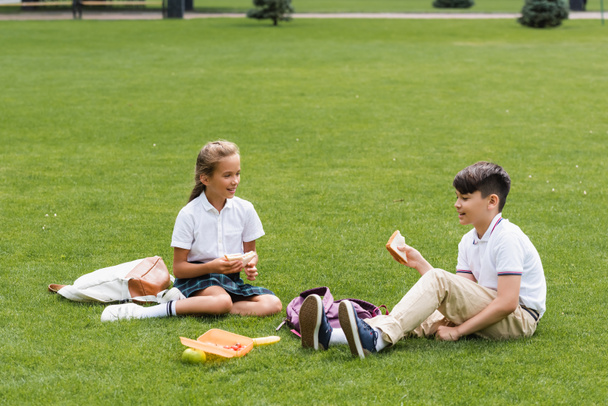 Wieloetniczni uczniowie trzymający kanapki w pobliżu plecaków na trawie w parku  - Zdjęcie, obraz