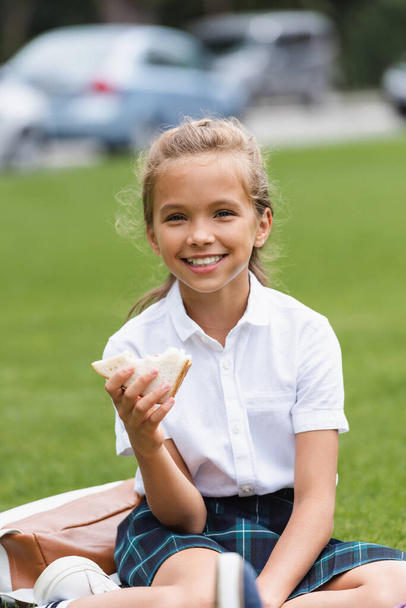 Smiling preteen schoolgirl holding sandwich on lawn in park  - Fotografie, Obrázek