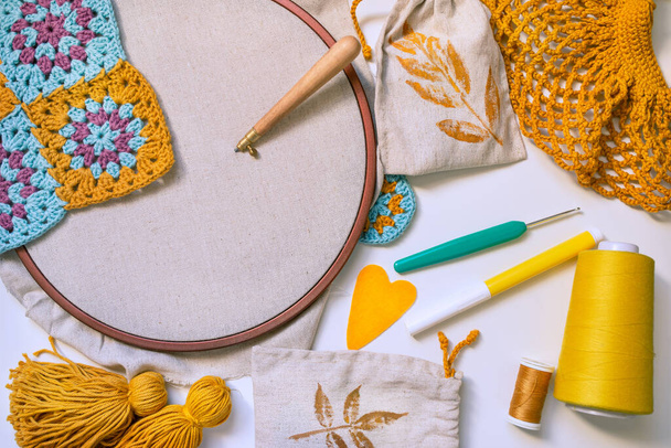 Flatlay of embroidery hoop with blank fabric and punch needle inserted in, crochet hook, tassels, heart, spool of thread, crochet patterns on white surface. Concept of creative hobby in yellow colors. - Photo, Image