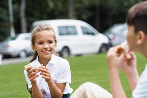 Okulda gülümseyen kız parkta bulanık sınıf arkadaşının yanında sandviç tutuyor.  - Fotoğraf, Görsel