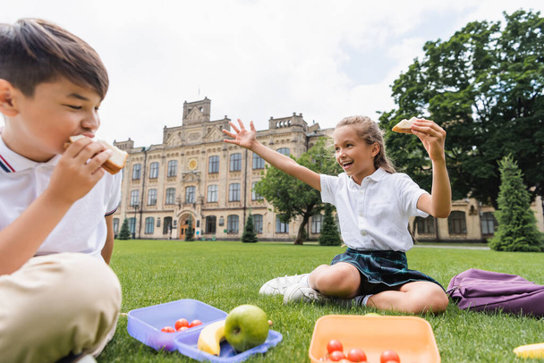 Schoolgirl holding sandwich and waving hand near lunchboxes and blurred asian friend on grass  - Photo, image