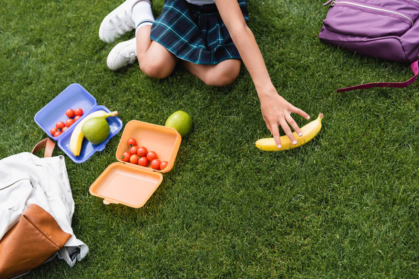 Vista recortada de colegiala tomando plátano cerca de mochilas y loncheras en la hierba  - Foto, Imagen