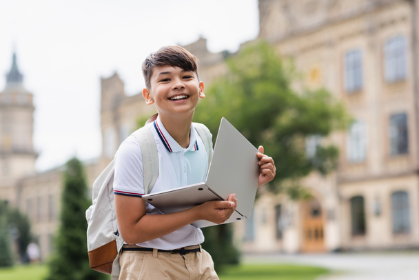 Cheerful asian schoolkid holding laptop and looking at camera outdoors  - Photo, Image
