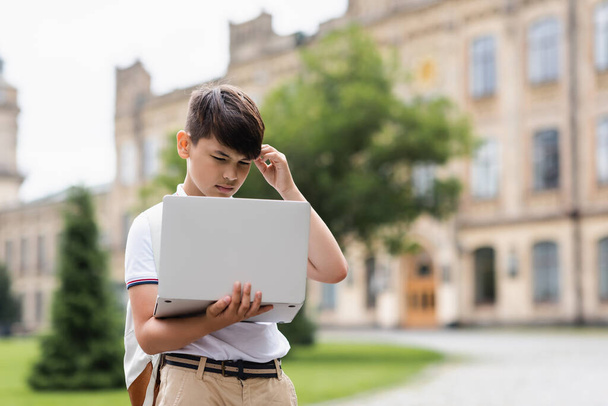 Pensive asian schoolboy looking at laptop outdoors  - Foto, imagen