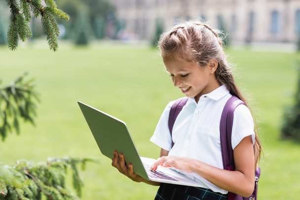 Smiling schoolgirl with backpack using laptop outdoors  - 写真・画像