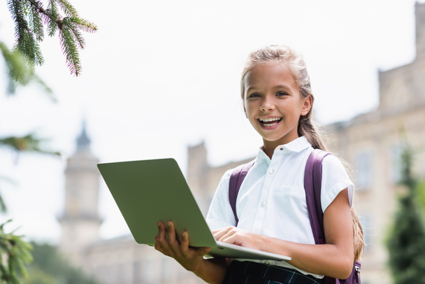 Happy schoolkid holding laptop and looking at camera outdoors  - Foto, Imagem