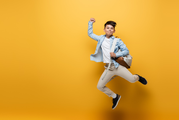 Asian schoolkid holding backpack and showing yes gesture while jumping on yellow background - Fotografie, Obrázek