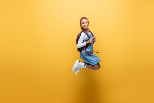 Cheerful schoolgirl with backpack jumping on yellow background - Foto, Imagem