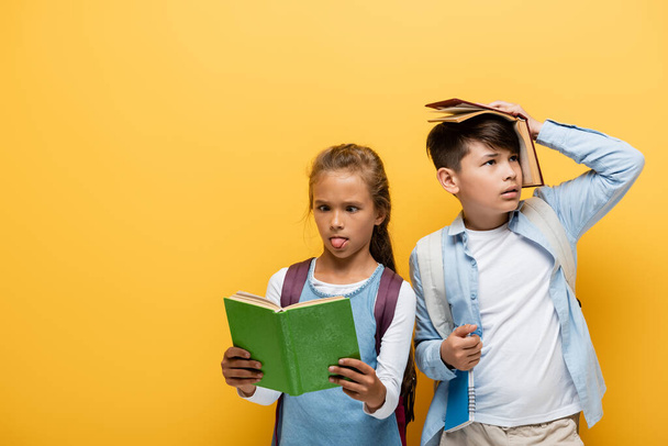 Écolière lecture livre et collant sur la langue près asiatique ami isolé sur jaune  - Photo, image