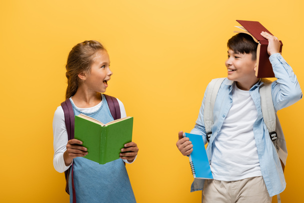 Pozytywne międzyrasowe dzieci trzymające książki i patrzące na siebie odizolowane na żółto  - Zdjęcie, obraz