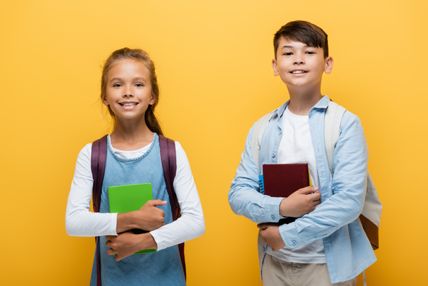 Cheerful multiethnic preteen schoolkids holding books and looking at camera isolated on yellow  - Foto, Imagem