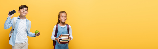 Des écoliers multiethniques joyeux tenant des livres et un téléphone cellulaire isolés sur du jaune, bannière  - Photo, image