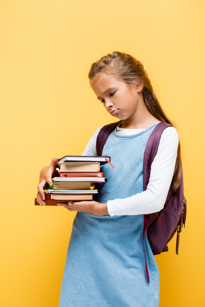 Upset preteen schoolgirl looking at books isolated on yellow  - Foto, imagen