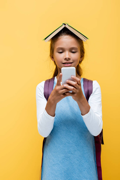 Smiling schoolgirl with book on head using mobile phone isolated on yellow  - Zdjęcie, obraz