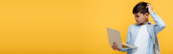 Pensive asian pupil with backpack looking at laptop isolated on yellow, banner  - Fotografie, Obrázek