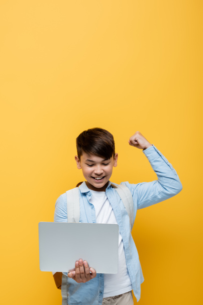 Aufgeregt asiatische Schulkind zeigt ja Geste und hält Laptop isoliert auf gelb  - Foto, Bild