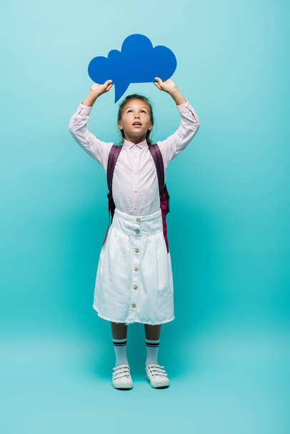 Full length of schoolgirl holding thought bubble on blue background  - Valokuva, kuva