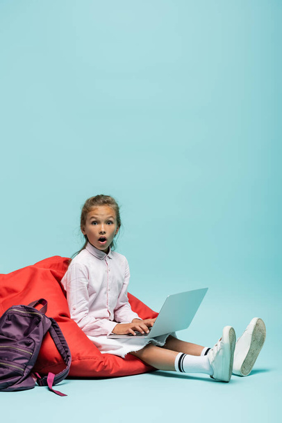 Geschokt schoolmeisje met behulp van laptop op zitzak stoel op blauwe achtergrond - Foto, afbeelding